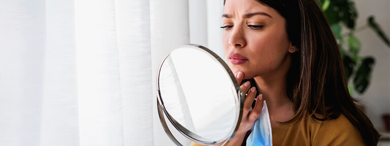 A concerned young woman examines her chin blemishes in a small circular mirror.