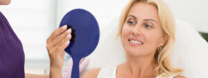 A satisfied woman looking at her reflection in a handheld mirror, after a laser face lift, with a medical professional standing beside her.